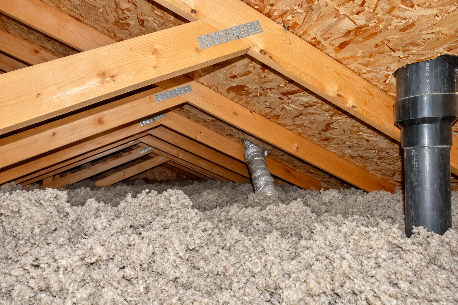 Gray loose-fill insulation blankets an attic floor beneath exposed roof trusses and a black pipe.