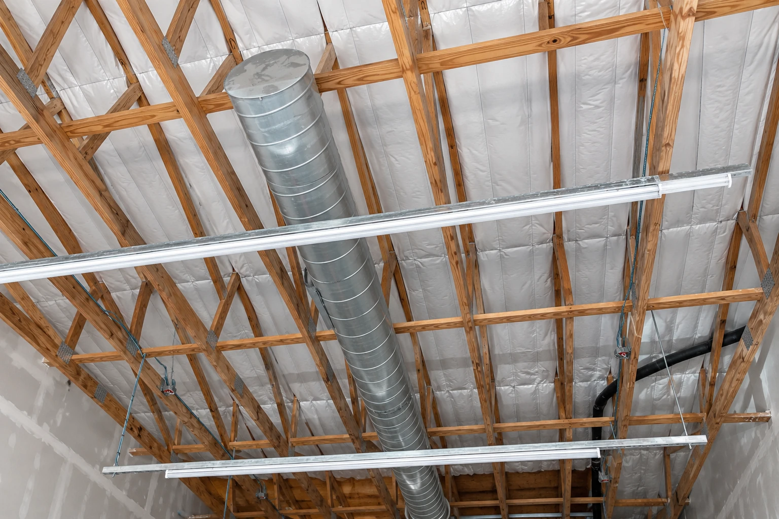 Commercial ceiling with white insulation between exposed wooden roof trusses, metal ductwork, and suspended light fixtures.