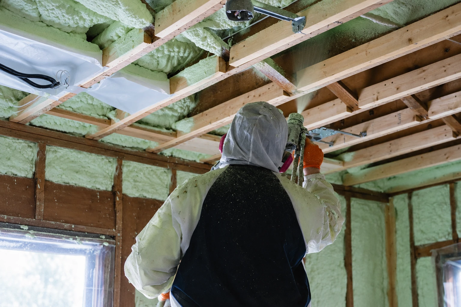 A worker in protective gear sprays foam insulation into the exposed ceiling and wall framing of a house.
