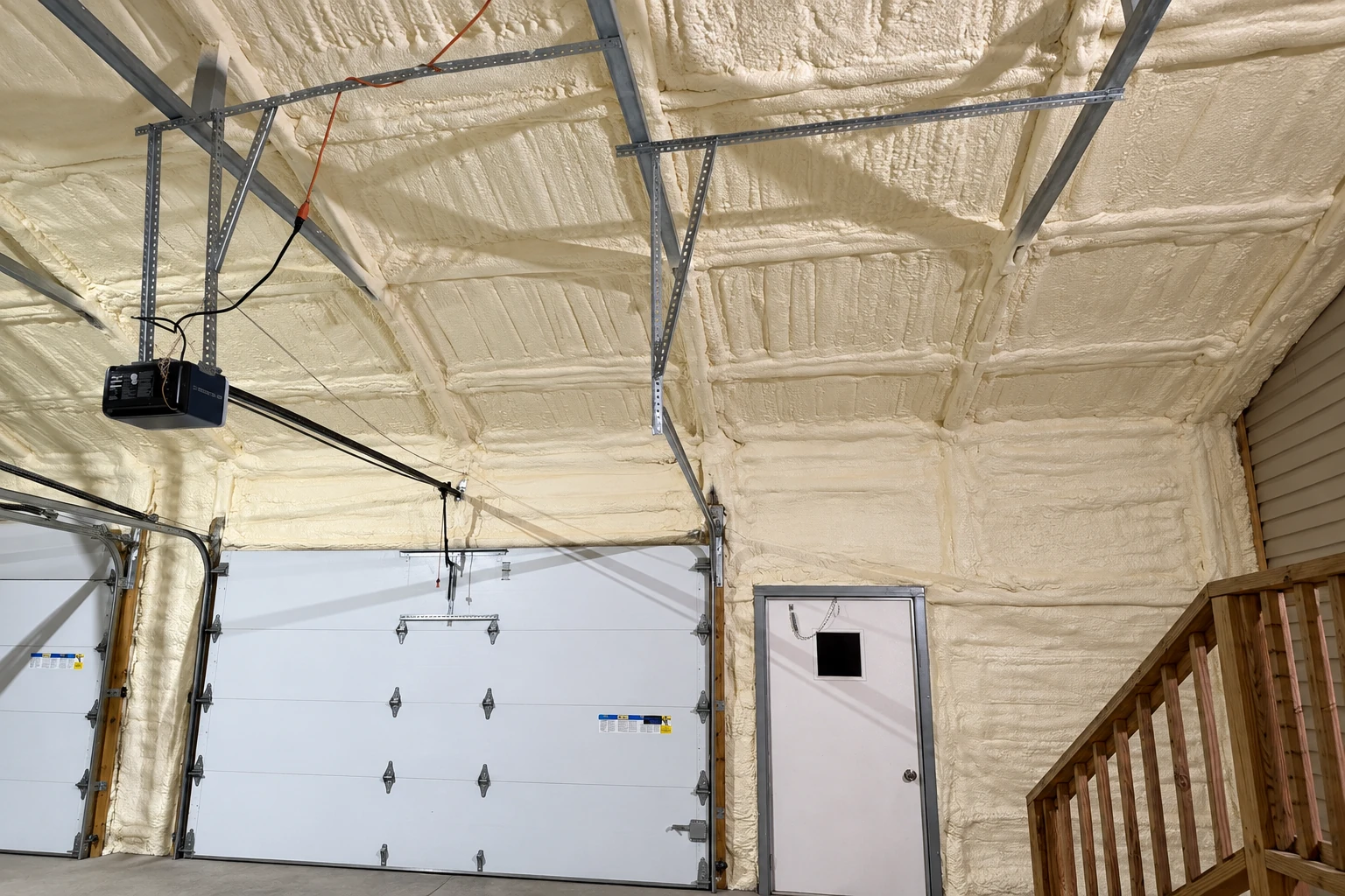 Cream spray foam covers the ceiling and walls of a garage around the doors, tracks, and overhead hardware.