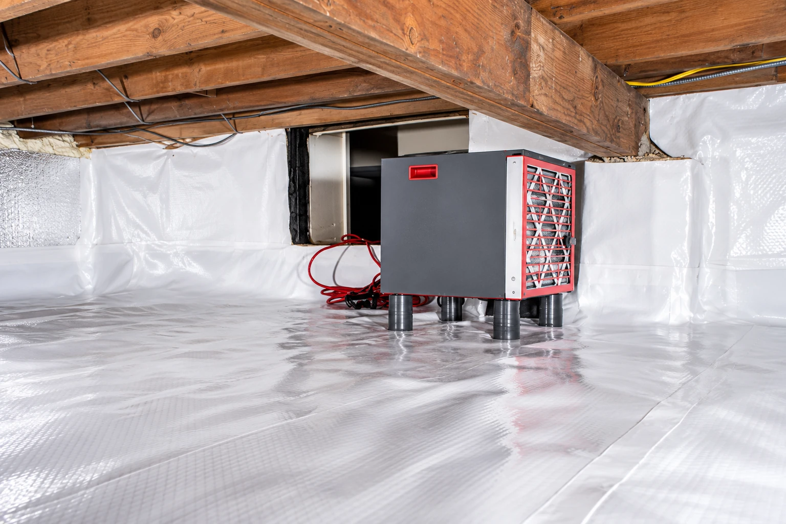White vapor barrier sheeting covers a crawl-space floor and walls around a gray dehumidifier beneath exposed wooden beams.