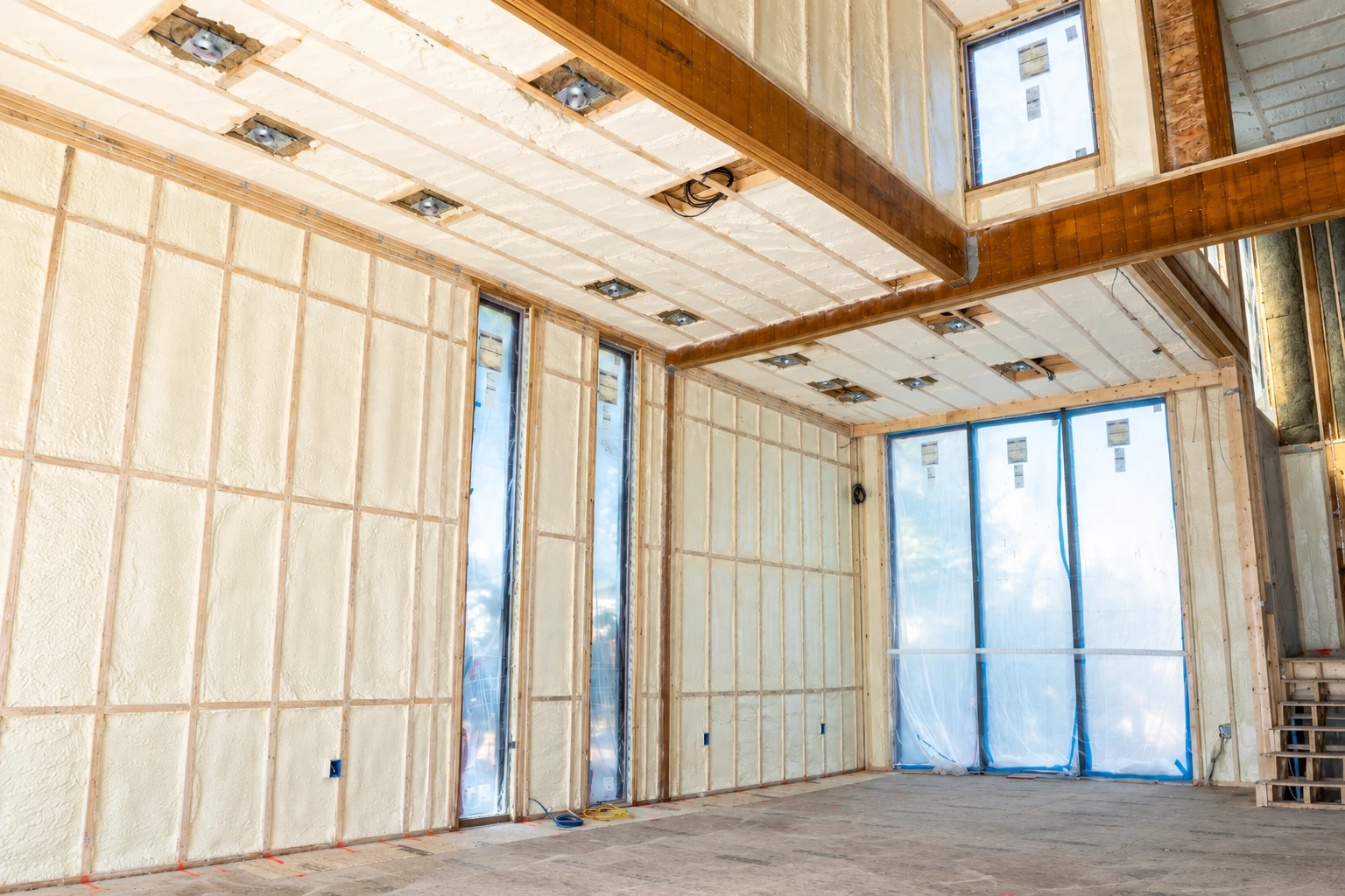 Unfinished room with pale spray foam insulation filling wall and ceiling framing around plastic-covered windows.